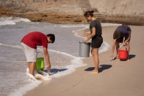 Vue de face du groupe de bénévoles multi-ethniques nettoyant la plage avec des seaux dans leurs mains par une journée ensoleillée — Photo de stock