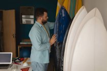 Vista lateral do homem caucasiano verificando e organizando pranchas de surf em uma oficina — Fotografia de Stock