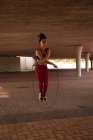 Вид спереди на молодую смешанную расовую женщину, тренирующуюся с скакалкой под мостом в городе — стоковое фото