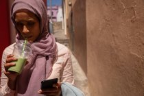 Vista frontal da bela mulher de raça mista usando telefone celular enquanto bebe um smoothie em um dia ensolarado — Fotografia de Stock