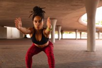 Vue de face de la jeune femme de race mixte faisant de l'exercice de squat sous un pont dans la ville — Photo de stock
