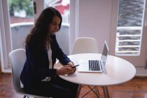 Seitenansicht einer schönen Frau mit braunem Haar, die ein Mobiltelefon benutzt, während sie im Büro an einem Laptop arbeitet — Stockfoto