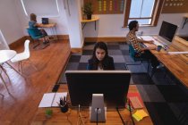 Vue de face d'une femme d'affaires métisse attentive travaillant sur ordinateur au bureau alors que ses collègues travaillent derrière elle au bureau — Photo de stock