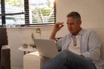 Vista frontale dell'uomo maturo caucasico che utilizza il computer portatile mentre è seduto sul divano in soggiorno a casa — Foto stock