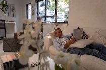 Vista frontal do homem caucasiano maduro usando laptop enquanto deitado no sofá na sala de estar em casa — Fotografia de Stock