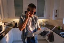 Vista frontal do homem maduro de pé e falando no telefone celular enquanto usa laptop na cozinha em casa — Fotografia de Stock