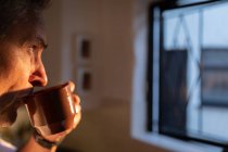 Side view of thoughtful mature Caucasian man having coffee and looking through the window in kitchen at home on sunrise — Stock Photo
