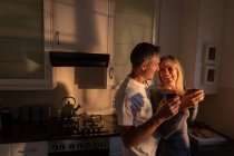 Side view of happy mature Caucasian couple having coffee while they are looking at each other in kitchen at home on sunrise — Stock Photo