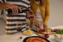Section médiane de l'homme verser du vin dans le verre tandis que la femme coupe des légumes et la préparation de pizza dans la cuisine à la maison — Photo de stock