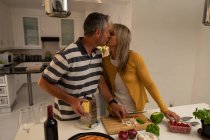 Vista frontal do casal maduro caucasiano se beijando enquanto prepara pizza na cozinha em casa — Fotografia de Stock