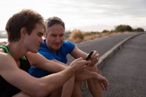Vista laterale del padre e del figlio caucasici guardando il telefono cellulare mentre si rilassano sulla strada — Foto stock