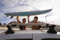 Вид спереди на Кавказского отца и сына, снимающих доску для сёрфинга с автомобильного носителя на пляже — стоковое фото