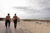 Vue arrière du père et du fils caucasiens marchant avec planche de surf à la plage — Photo de stock