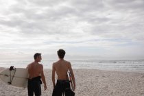 Vista posteriore di padre e figlio caucasico in piedi con tavola da surf in spiaggia — Foto stock