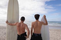 Вид сзади на Кавказских отца и сына, стоящих с доской для серфинга на пляже — стоковое фото