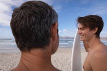 Vista posteriore di padre e figlio caucasico con tavola da surf interagire tra loro in spiaggia — Foto stock