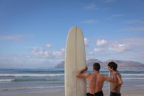 Visão traseira do pai e do filho caucasianos de pé com prancha de surf na praia em um dia ensolarado — Fotografia de Stock