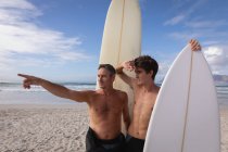 Vue de face du père et du fils caucasiens avec planche de surf interagissent les uns avec les autres à la plage — Photo de stock