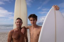 Retrato de pai e filho caucasiano em pé com prancha de surf na praia — Fotografia de Stock