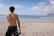 Vista posteriore dell'uomo caucasico in piedi con tavola da surf in spiaggia in una giornata di sole — Foto stock