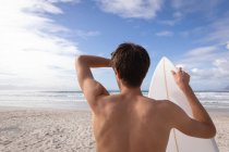 Rückansicht eines kaukasischen Mannes, der an einem sonnigen Tag mit Surfbrett am Strand steht — Stockfoto