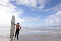 Visão traseira do homem caucasiano de pé com prancha de surf na praia em um dia ensolarado — Fotografia de Stock