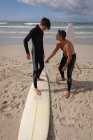 Vista frontal do pai caucasiano assistir filho para montar prancha de surf na praia em um dia ensolarado — Fotografia de Stock