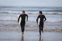 Vista frontal de feliz padre e hijo caucásico con tabla de surf corriendo fuera del mar - foto de stock