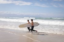 Вид сбоку на отца и сына кавказца с доской для серфинга, выбегающей из моря в солнечный день — стоковое фото