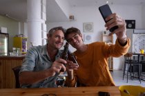 Vista frontal del padre y el hijo caucásicos tomando selfie desde el teléfono móvil mientras brindan cerveza en casa - foto de stock