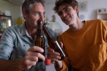 Primer plano de padre e hijo caucásicos tostando botella de cerveza en casa - foto de stock