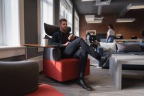 Vista lateral do empresário caucasiano olhando para seu smartwatch enquanto sentado no sofá no escritório — Fotografia de Stock