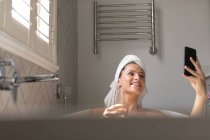 Femme souriante avec verre de champagne prenant un selfie dans la baignoire dans la salle de bain — Photo de stock
