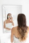 Вид спереду жінки, що дивиться у дзеркало і наносить маску для обличчя після ванни — стокове фото