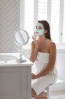 Mulher olhando no espelho e aplicando máscara facial após o banho no banheiro — Fotografia de Stock