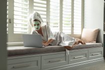 Femme en peignoir couchée et utilisant un ordinateur portable près de la fenêtre à la maison — Photo de stock