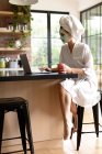 Жінка з чашкою кави використовує ноутбук на кухні після ванни вранці — стокове фото