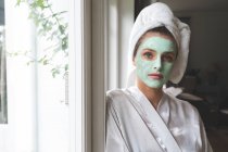 Beautiful woman in bathrobe wearing facial mask, leaning against the window — Stock Photo