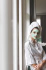 Schöne Frau im Bademantel mit Gesichtsmaske, gegen das Fenster gelehnt — Stockfoto