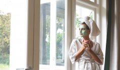 Beautiful woman in bathrobe wearing facial mask, looking through the window — Stock Photo