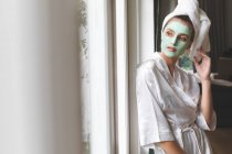 Belle femme en peignoir portant un masque facial, regardant par la fenêtre — Stock Photo