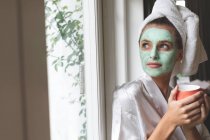 Mulher bonita em roupão de banho usando máscara facial, olhando pela janela — Fotografia de Stock