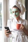 Красива жінка в масці для обличчя, використовуючи мобільний телефон біля вікна вдома — стокове фото
