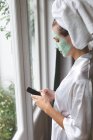 Красивая женщина в маске с помощью мобильного телефона возле окна дома — стоковое фото
