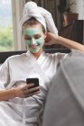 Вид спереду жінки в масці для обличчя з використанням мобільного телефону на дивані вдома — стокове фото