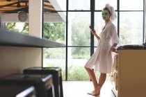 Вид збоку жінки в масці для обличчя, використовуючи мобільний телефон на кухні вдома — стокове фото