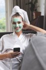 Vista frontale della donna in maschera facciale utilizzando il telefono cellulare sul divano a casa — Foto stock