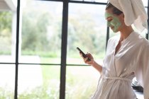 Вид сбоку женщины в маске на лице с помощью мобильного телефона на кухне дома — стоковое фото