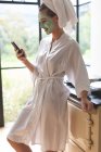 Seitenansicht einer Frau mit Gesichtsmaske mit Handy in der heimischen Küche — Stockfoto