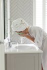 Mujer vista lateral lavándose la mascarilla en el lavabo del baño en casa - foto de stock